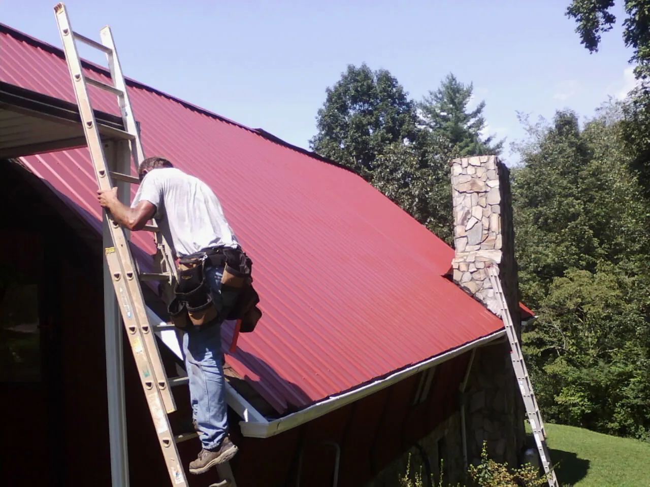 Roofer on a ladder beside a red metal roof and stone chimney — Soffit & Fascia Repair by Roofing Brothers in Stratton
